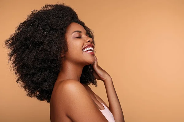 Side view of merry African American woman with curly hair touching clean face and laughing with closed eyes during spa session against brown background.