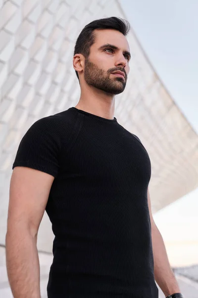 From below young Middle Eastern male in black t shirt looking away while standing against roof of contemporary building on city street