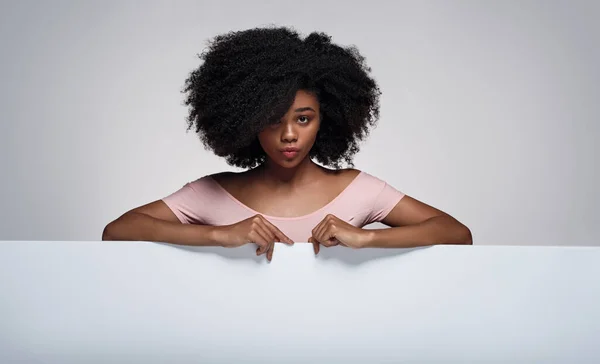 Афроамериканка Вьющимися Волосами Смотрит Камеру Дуется Губами Указывая Пустой Плакат — стоковое фото