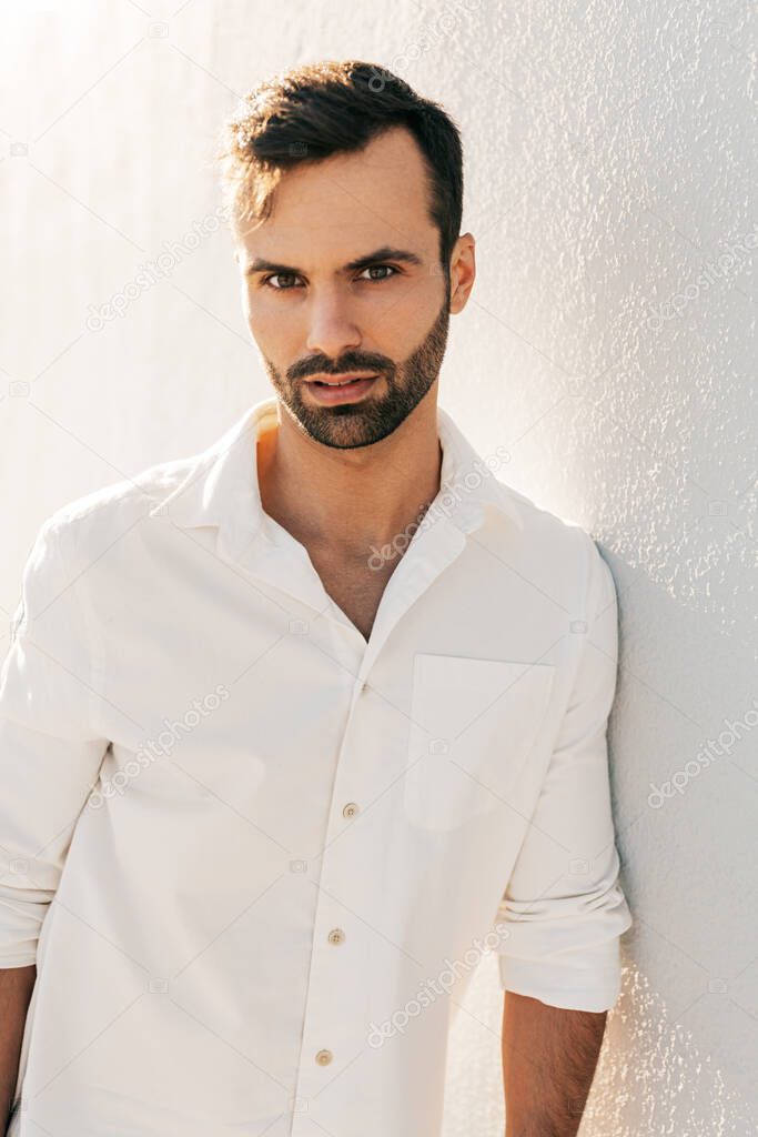 Self assured young bearded Arab male with dark hair in white shirt leaning on wall and looking at camera, while standing on street at sunset