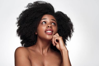 Çıplak omuzlu, kıvırcık saçlı, yüzüne dokunan ve yukarı bakan genç bir Afro-Amerikan kadın model. Gri arka planda cilt bakımı prosedürünü düşünürken tereddüt içinde.