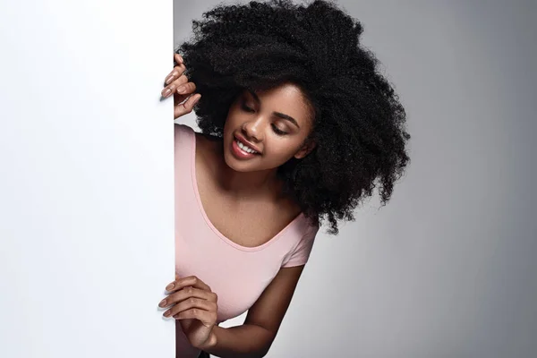 Cheerful young African American lady with curly dark hair in casual clothes smiling and peeking behind white poster against gray background in studio