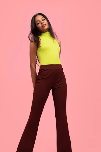 Low angle of confident young Latin American female millennial with long dark hair, in trendy outfit standing against pink background with hands behind back and looking at camera