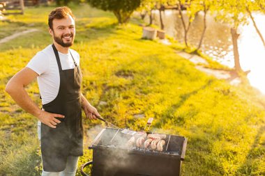 Beyaz tişörtlü ve siyah önlüklü sakallı erkek şef, yaz günü gölün kıyısındaki barbekü sırasında ızgarada biftek ve sosis pişirirken kameraya bakıyor.