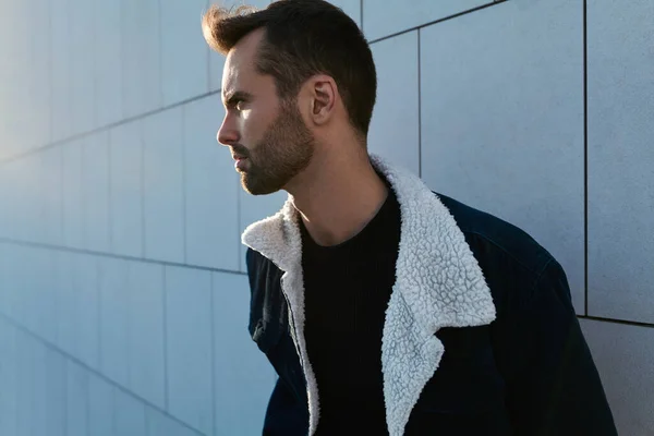 Concentrated young bearded ethnic male with dark hair in stylish casual clothes standing near tiled wall and looking away on sunny day in city