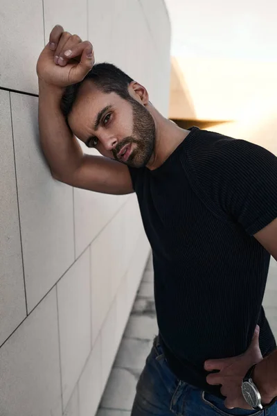 Self assured young bearded Middle Eastern male model with dark hair in black t shirt, leaning on wall and looking at camera while standing on street with hand on waist