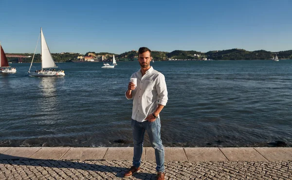 Self assured young ethnic male with dark hair and beard in casual outfit drinking takeaway, coffee and looking at camera while standing on paved embankment near lake on sunny day