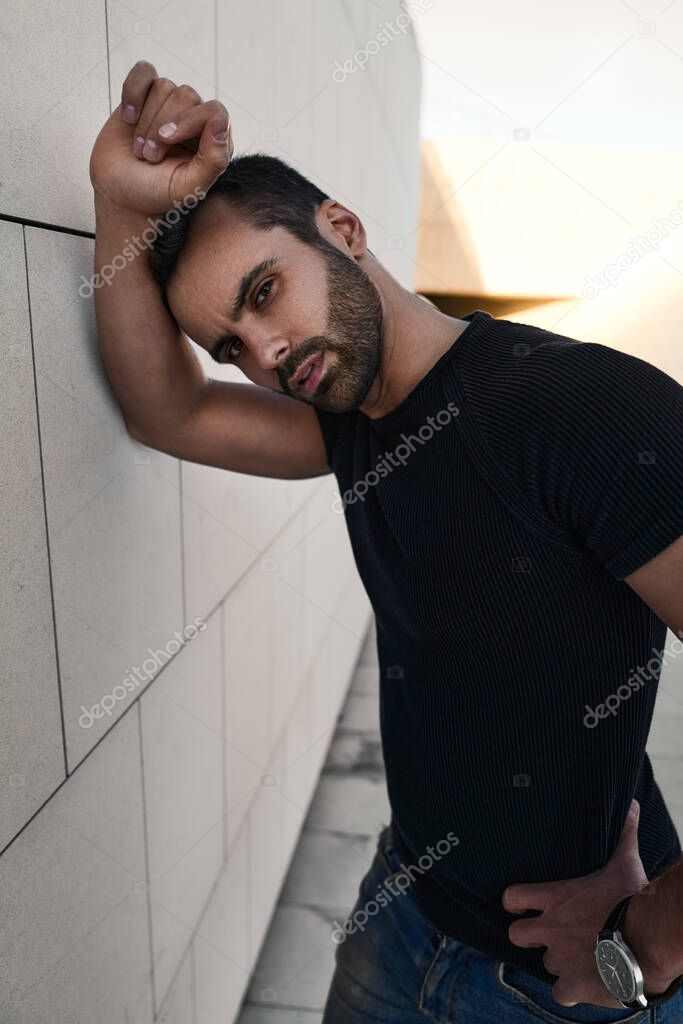 Self assured young bearded Middle Eastern male model with dark hair in black t shirt, leaning on wall and looking at camera while standing on street with hand on waist