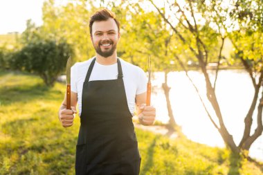 Çatal ve bıçaklı arkadaş canlısı sakallı adam, güneşli bir yaz günü, barbekü sırasında gölün kıyısında durup kameraya bakıp gülümsüyor.