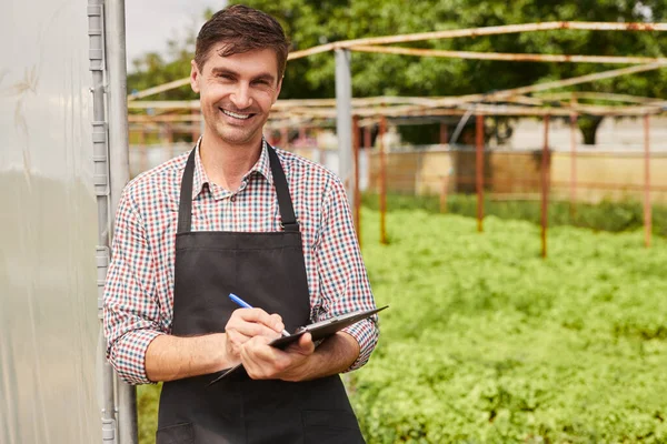 Cheerful adult man in checkered shirt and black apron looking at camera with smile and writing on clipboard, while standing near field of fresh lettuce during work on farm in summer