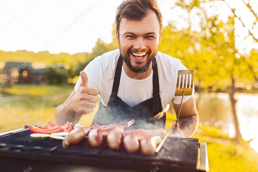 Optimistic bearded male chef with spatula looking at camera with smile and gesturing thumb up, while grilling sausages and vegetables during picnic on lake shore in summer weekend