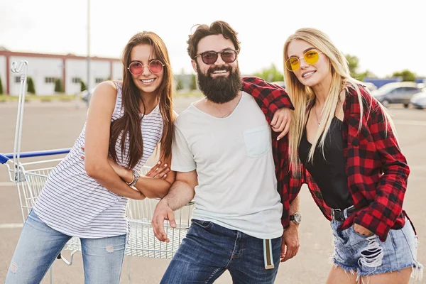 ショッピングカートの近くの駐車場に立っている間 カジュアルな服やサングラスで陽気な若い多人種の男性と女性の友人 笑顔とカメラを見て — ストック写真