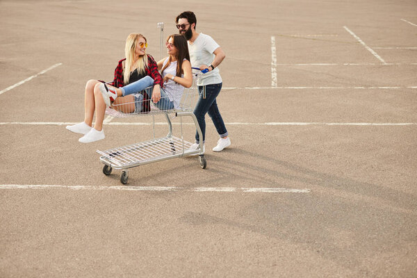 Бородатый хипстер в повседневной одежде и солнцезащитных очках толкает тележку с молодыми женщинами во время прогулки по парковке во время шоппинга в выходные дни