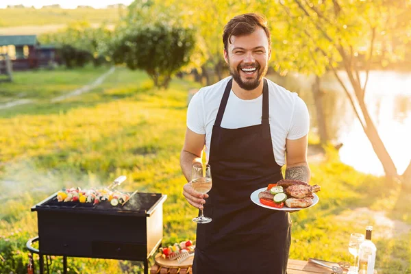 Joyful young bearded guy in t shirt and apron laughing and looking at camera, while standing on grassy lawn near lake with plate of grilled meat and veggies and wineglass