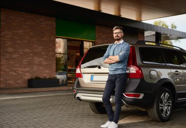 Sıradan giyinmiş, gözlüklü, kollarını kavuşturmuş sakallı bir adam şehrin benzin istasyonunda modern bir araca yaslanırken kameraya bakıyor.