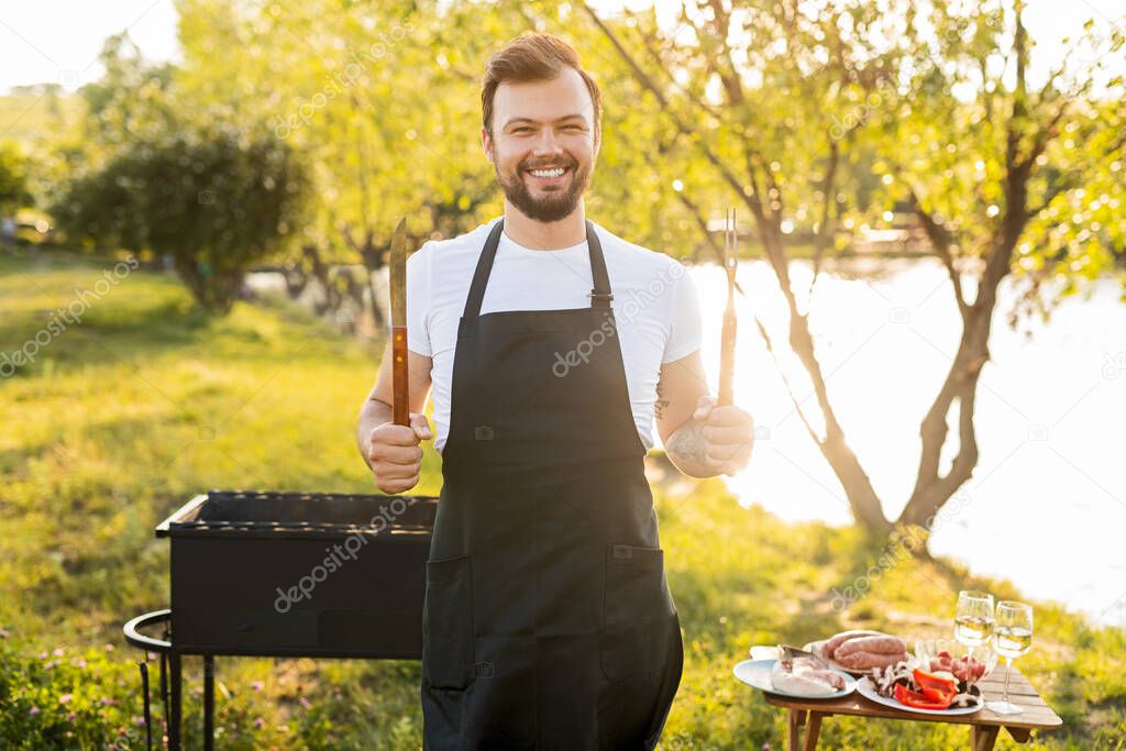 Happy bearded man in white t shirt and black apron with knife and fork smiling, and looking at camera during picnic on lake shore on sunny summer day