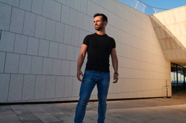 Güneşli bir günde çağdaş binanın yanında asfalt bir caddede dikilirken, sırıtan ve başka yöne bakan, siyah saçlı, kendine güvenen genç ve etnik bir erkekten.