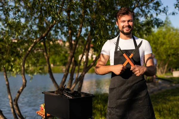 Man in white t shirt and black apron crossing fork and knife and looking at camera with smile, while standing near grill during barbecue on lake coast in daytime