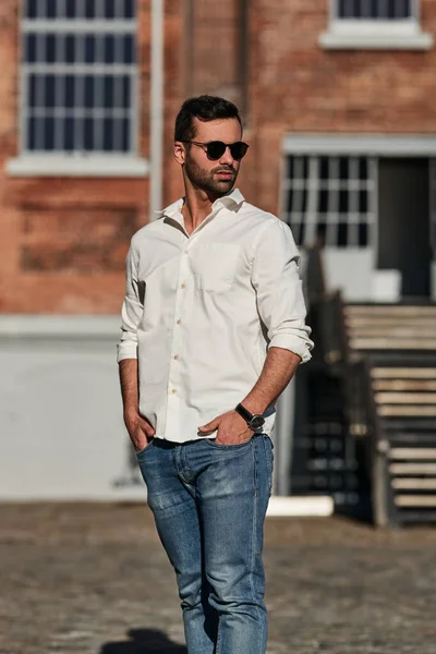 Self assured ethnic male in jeans and white shirt with stylish sunglasses holding hands in pockets and looking away, while standing on sunlit city street outside brick building