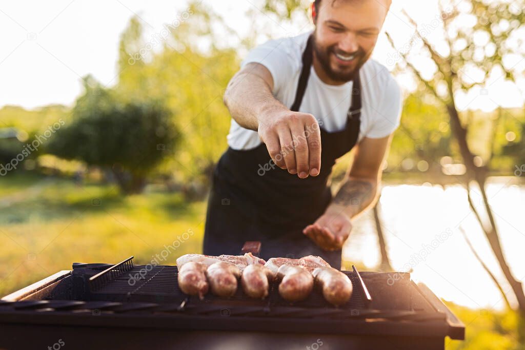 Soft focus of happy male chef in apron smiling and adding salt to sausages and meat on grill, while preparing food for picnic on summer day in countryside