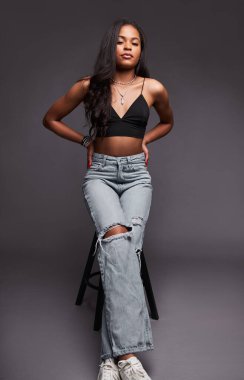Young Latin American female in jeans and bra holding hands on waist and looking at camera while leaning on stool against gray background