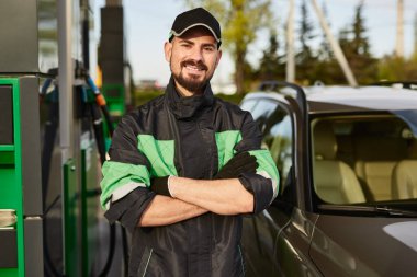 Positive young bearded male worker in uniform and cap smiling at looking at camera, while standing at gas station with crossed arms on sunny day