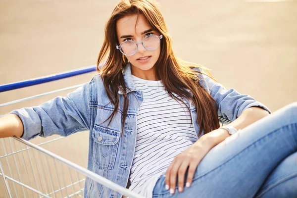 Self assured young female millennial with long brown hair in casual outfit and trendy, eyeglasses sitting in shopping cart and looking at camera on street on sunny day