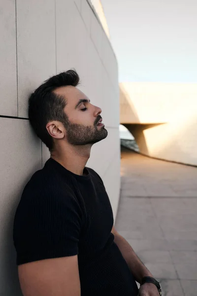 Bearded ethnic male in black t shirt closing eyes and leaning on tiled wall of sunlit building on city street in daytime