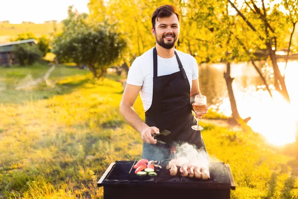 Positive bearded male chef in black apron with glass of wine smiling and looking at camera, while preparing sausages and vegetables on grill during barbecue on sunny summer day on lake coast