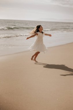 Beyaz elbiseli mutlu küçük kız kollarını açıyor ve etrafta dönüyor yazları dalgalı deniz kenarında kumlu sahilde eğlenirken