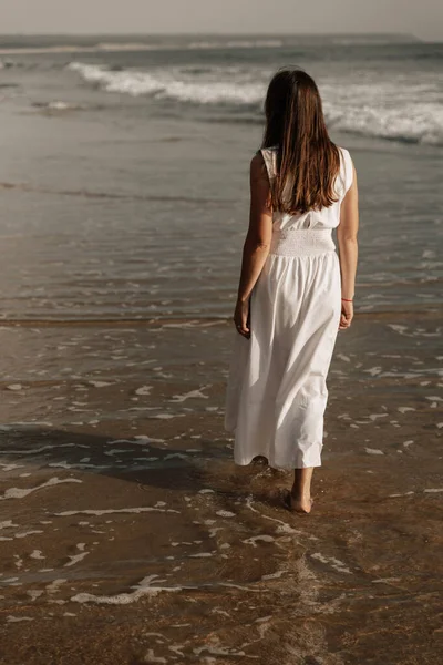 Back view of unrecognizable barefooted calm female tourist with long dark hair in white dress, walking on sandy coast washed by wavy ocean at sunset