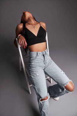 Kotlu ve sütyenli Latin Amerikalı kadın manken sandalyede oturuyor ve gri arka plana yaslanıyor.