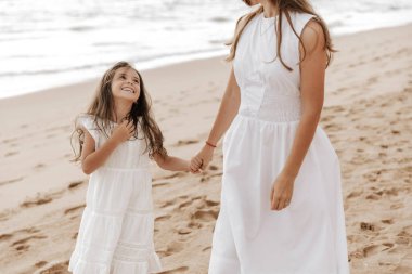 Beyaz elbiseli samimi bir çocuk. Mahsul annesinin elini tutuyor ve kumlu sahilde birlikte dolaşırken gülümsüyor.