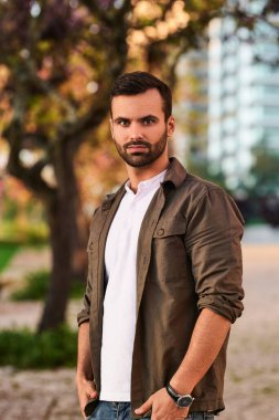 Kendine güvenen, siyah saçlı, şık giyimli genç erkek model. Şehir parkında, elleri ceplerinde ve kameraya bakıyor.