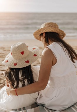 Tarz sahibi, beyaz elbiseli, saman şapkalı, uzun siyah saçlı, kumlu sahilde birlikte oturmuş, deniz manzarasına hayran olan sevimli küçük bir kızı kucaklayan tanınmamış bir anne.