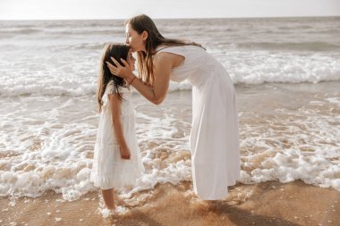 Uzun kahverengi saçlı, genç ve şık bir kadın, mutlu küçük bir kızın alnını öperken ıslak kumlu sahilde köpüklü okyanus tarafından yıkanırken.