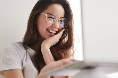Low angle of optimistic teen student in glasses touching face and smiling while browsing data on laptop