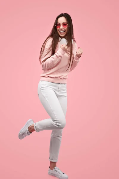 Full body happy teen brunette in casual clothes with sunglasses clenching fists and looking at camera with smile while celebrating victory against pink background