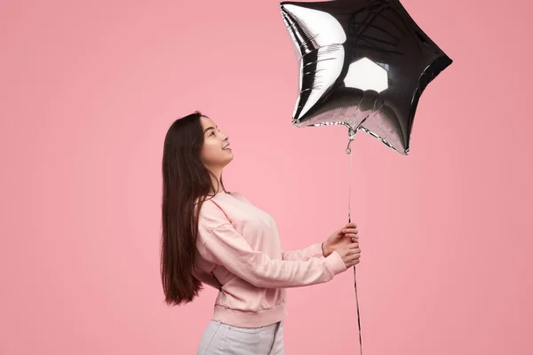 Side view of positive young female millennial with long dark hair in sweatshirt smiling, while holding silver star shaped balloon against pink background during birthday celebration