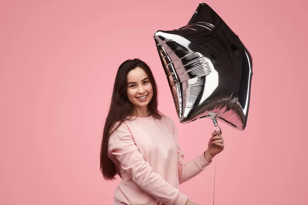 Delighted young female model with long dark hair in sweatshirt smiling happily, while holding silver star shaped balloon and looking at camera against pink background during birthday celebration