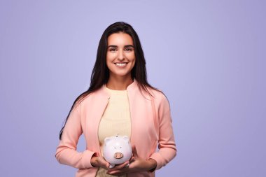 Happy young female with long dark hair holding piggy bank and looking at camera against violet background