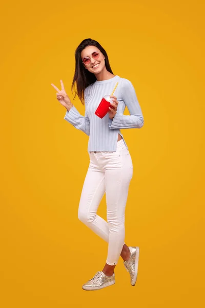 Full body happy young brunette in casual clothes with takeaway cup of milkshake gesturing V sign and looking at camera against yellow background