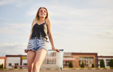 Low angle of glad blond woman in shorts and top with sunglasses smiling and looking away, while leaning on shopping trolley on blurred background of cloudy sky and mall