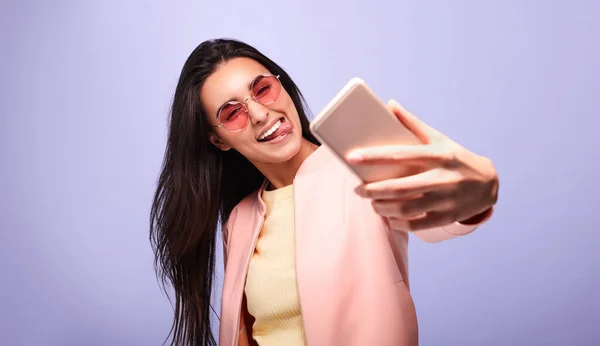Happy young female in trendy outfit and sunglasses smiling and taking selfie while standing against violet background
