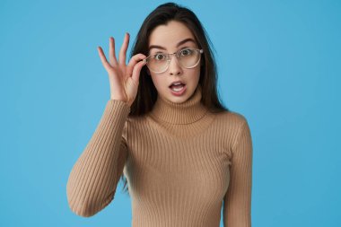 Uzun siyah saçlı, bej yakalı, gözlük takan ve mavi arka planda ağzı açık kameraya bakan hayret verici genç bayan öğrenci.