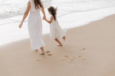 Yüksek açılı, sevimli, mutlu, yalınayak, beyaz elbiseli, uzun siyah saçlı küçük kız kumlu sahilde dalgalı okyanusa doğru yürüyor ve anonim bir annenin elini tutuyor.