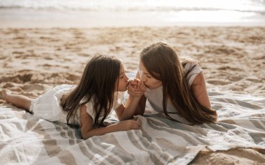Loving mom and daughter holding and kissing hands while lying on blanket on summer weekend day on sandy beach