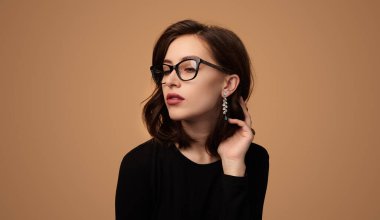 Zarif gözlüklü ve küpeli kendine güvenen genç bir bayan model koyu renk saçlara dokunuyor ve bej arka plana bakıyor.