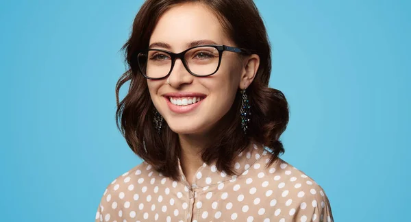 Clever young woman in dotted blouse and stylish glasses with dark hair smiling and looking away against blue background