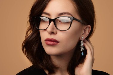 Crop confident young female model in stylish glasses and earrings touching dark hair and looking away against beige background